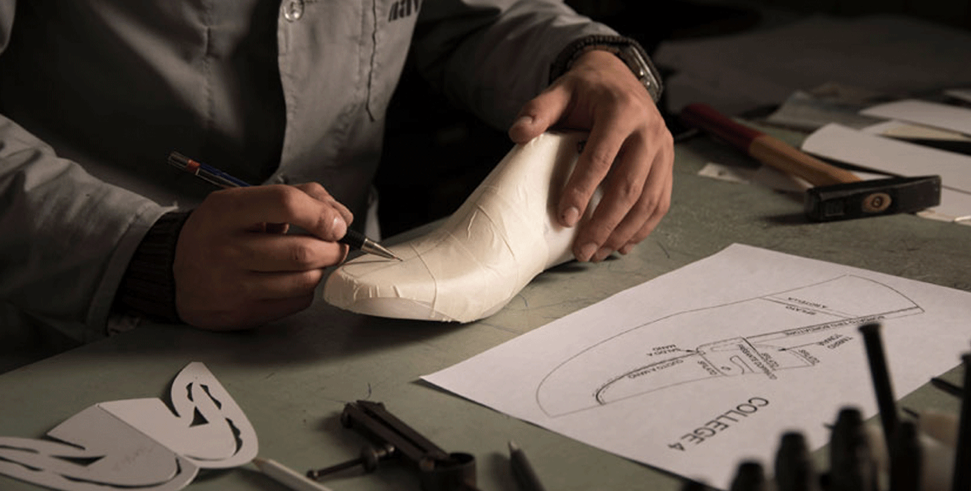 A maker drawing an upper pattern on a taped last beside paper patterns and tools